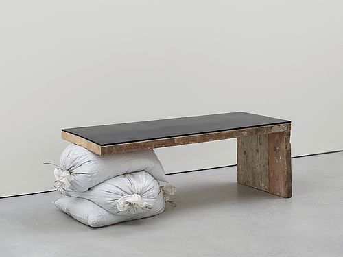 Eine Bank aus abgenutzten Holzbrettern, deren Sitzfläche mit schwarzem Gummi überzogen ist. Anstelle eines linken Beines dienen drei übereinandergestapelte weisse Säcke als Stütze. Die improvisierte Konstruktion und die gefundenen Materialien stehen im Kontrast zum sauberen, grauen Museumsinterieur.